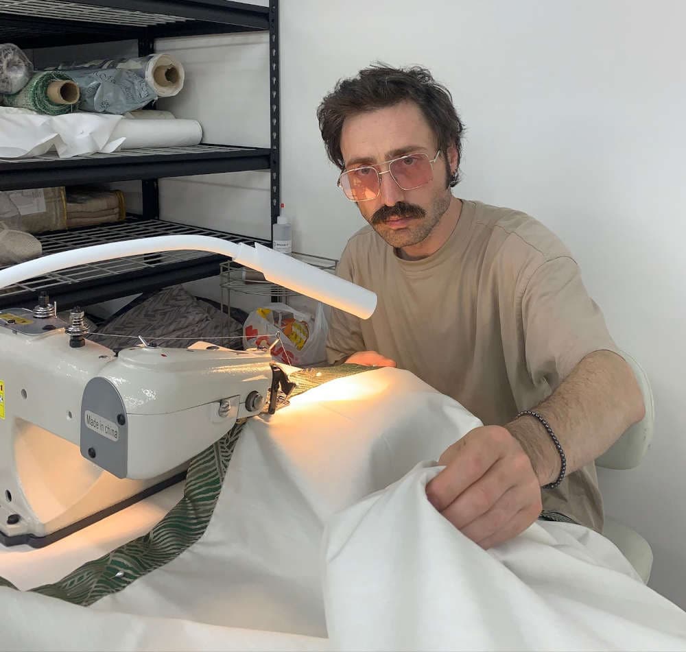 An employee at Emra Home Fashion sewing fabric on a machine, with fabric rolls in the background.