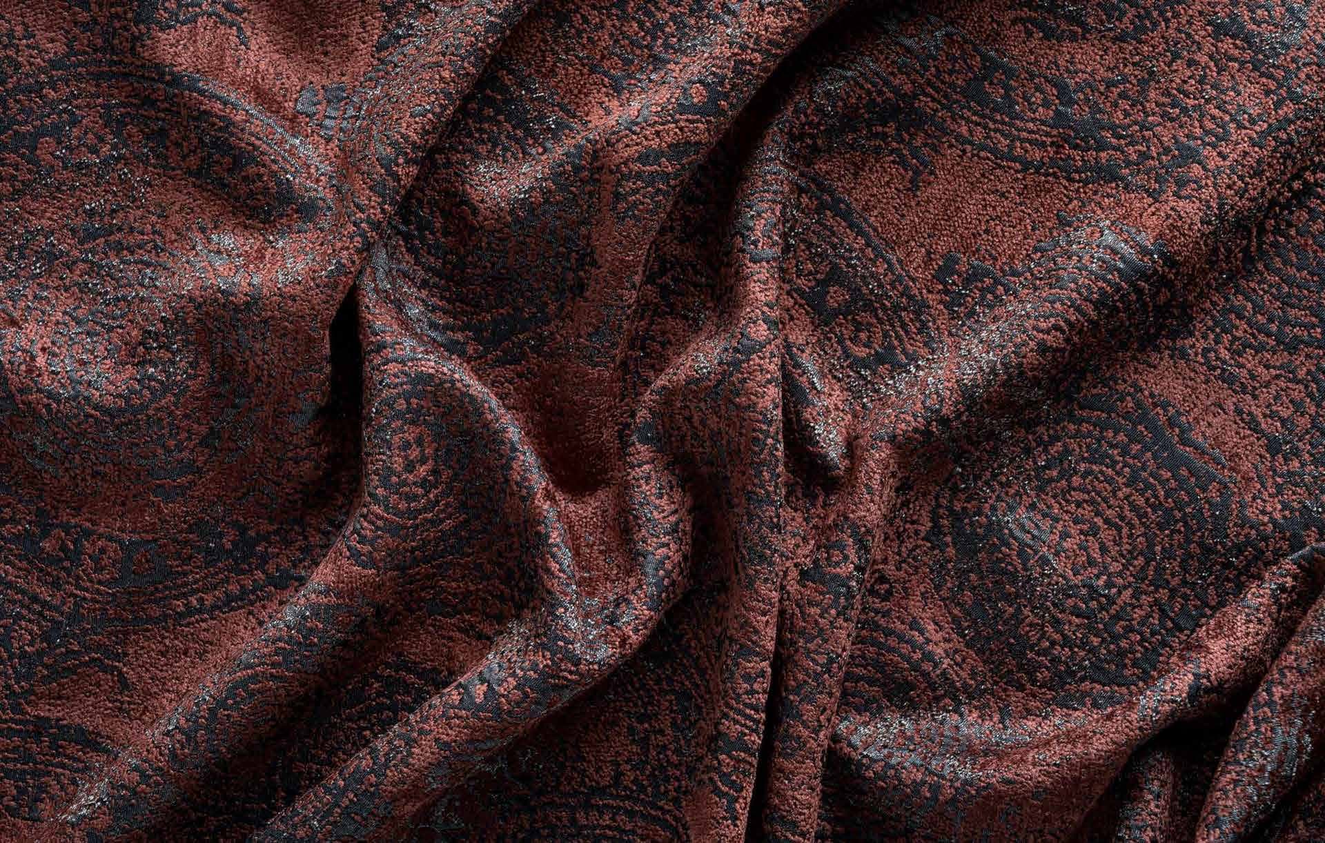 A close-up of textured fabric with an intricate pattern (possibly paisley or circular motifs) in shades of brown/copper and dark blue/black.
